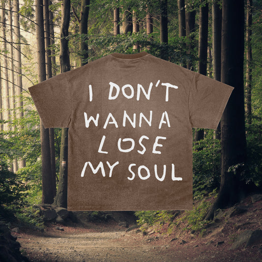 A brown crew neck t-shirt with the text 'I DON'T WANNA LOSE MY SOUL' printed on the front, displayed on a wooden hanger against a forest backdrop.